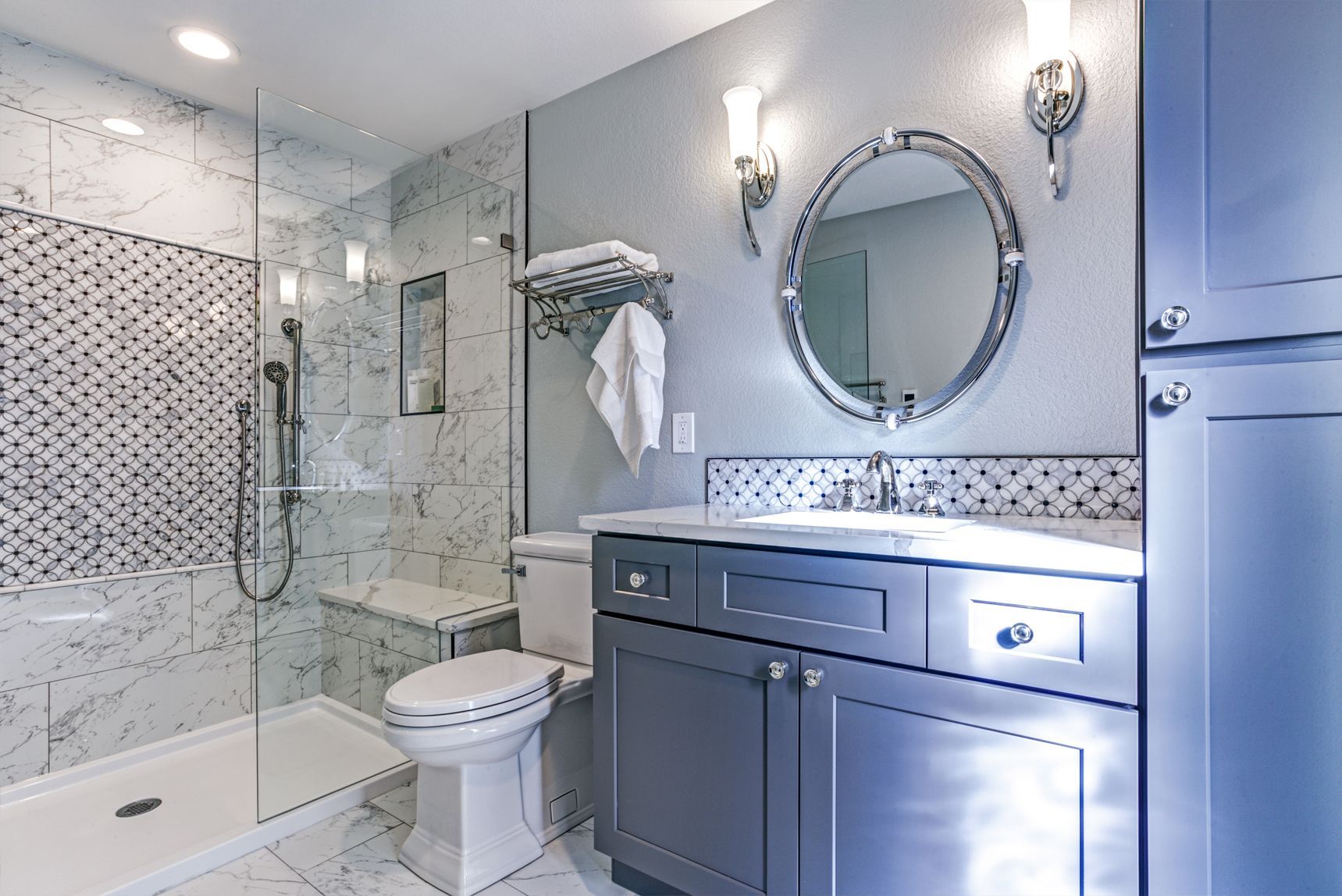 Modern bathroom with blue-grey cabinets, marble-like tile, shower, toilet, and vanity with a round mirror.