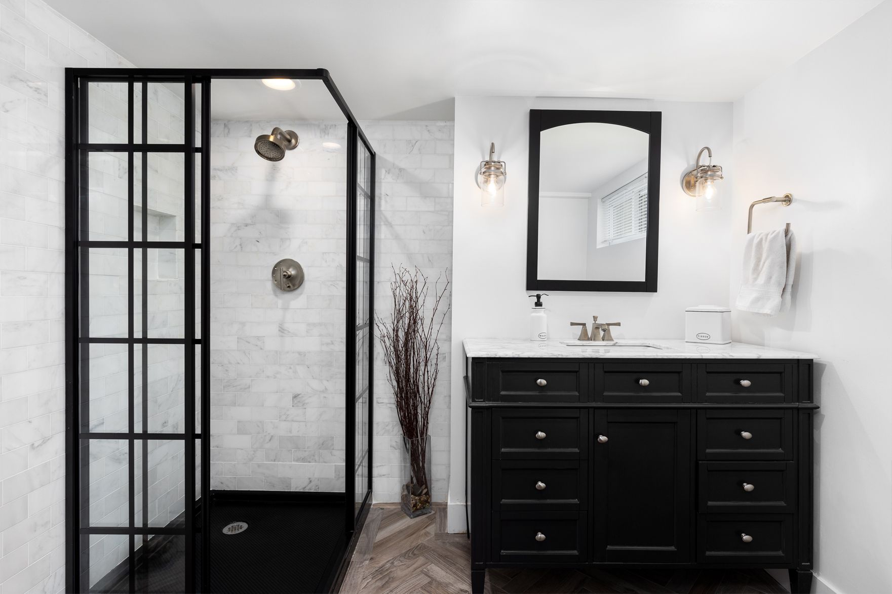 Black and white bathroom with a glass shower and black vanity; white walls, marble tiles, and hardwood floor.