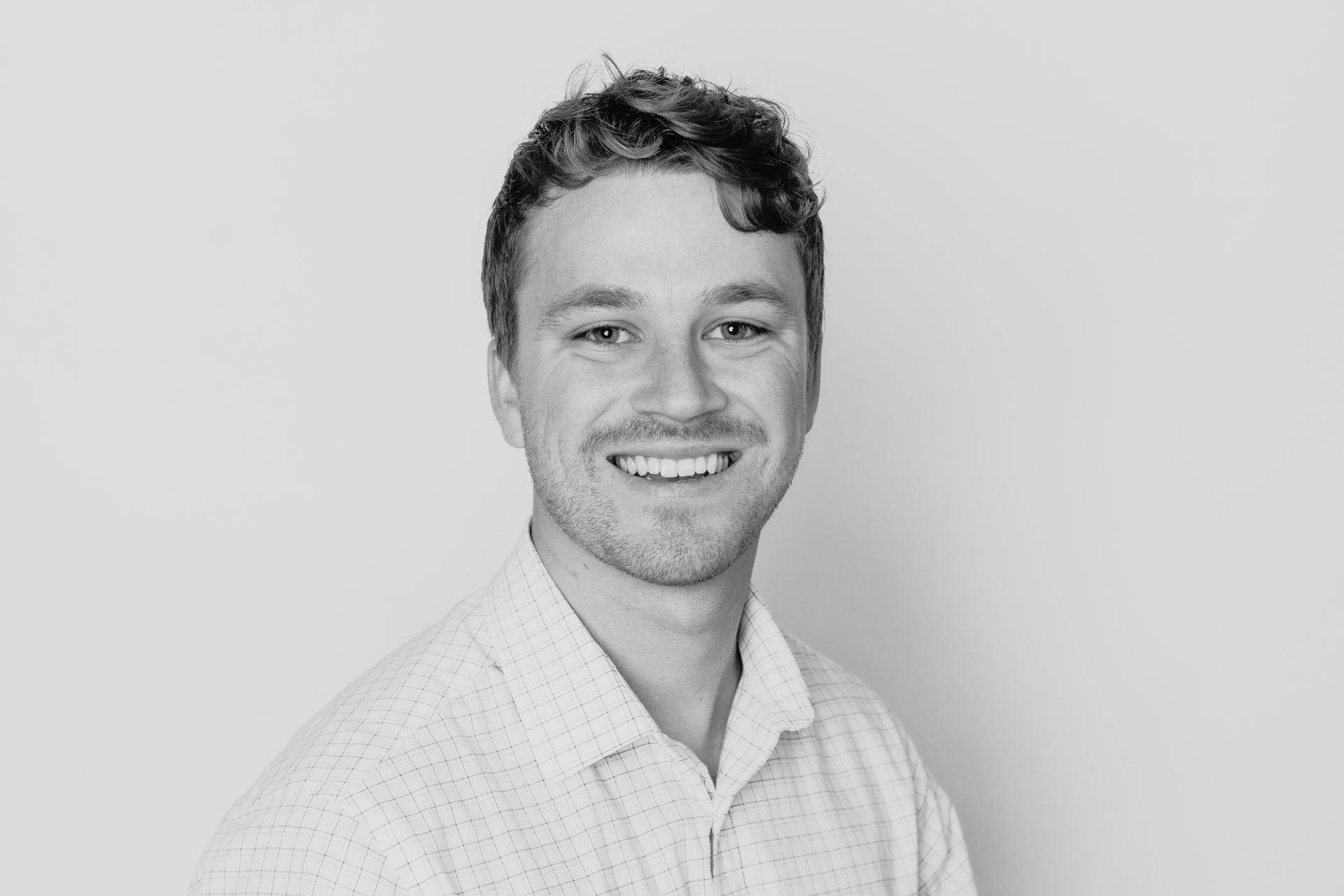 A person with short, wavy hair and a light-colored collared shirt smiling against a neutral gray background.