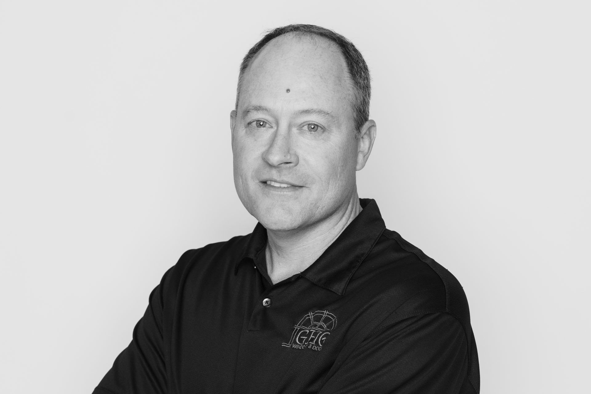 Headshot of a person in a black polo shirt with arms crossed, posing against a plain light gray background.