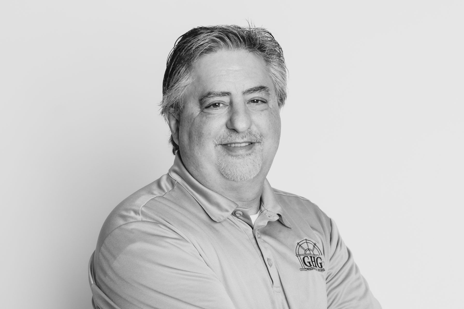 A headshot of a smiling person with graying hair wearing a collared polo shirt, posed with arms crossed against a backdrop.