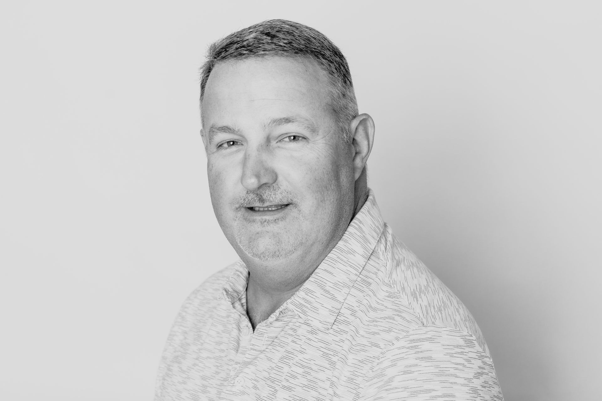 A man with short hair and a patterned button-down shirt smiling slightly against a plain, light-colored background.