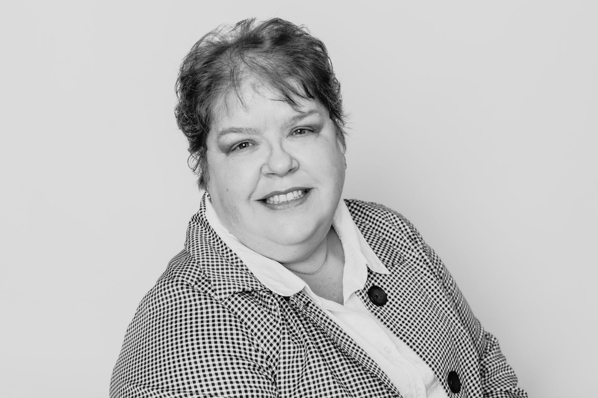 A person in a patterned blazer and white collared shirt smiles at the camera against a plain, light background.