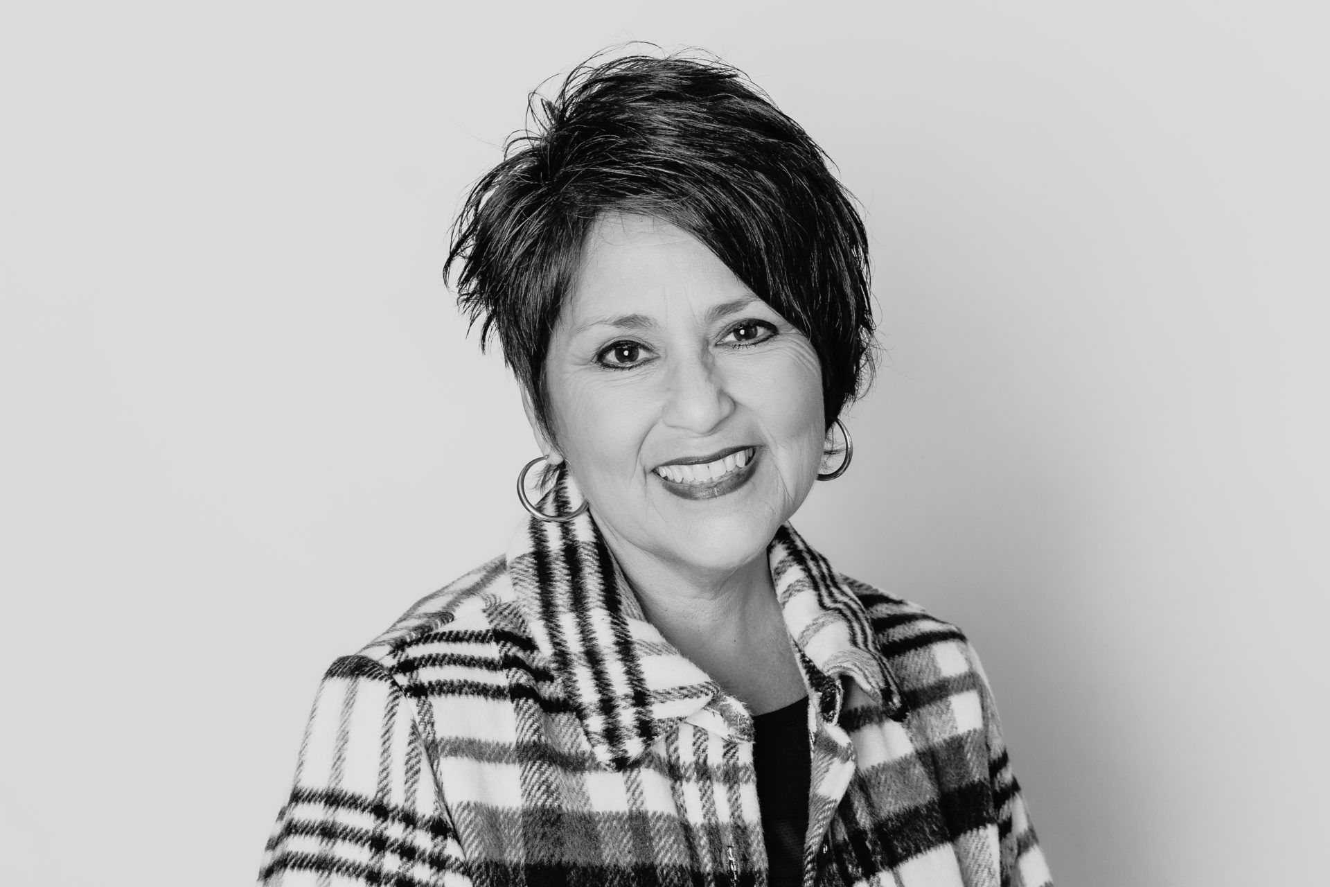 A smiling person with short, dark hair wearing a patterned plaid jacket against a plain background.