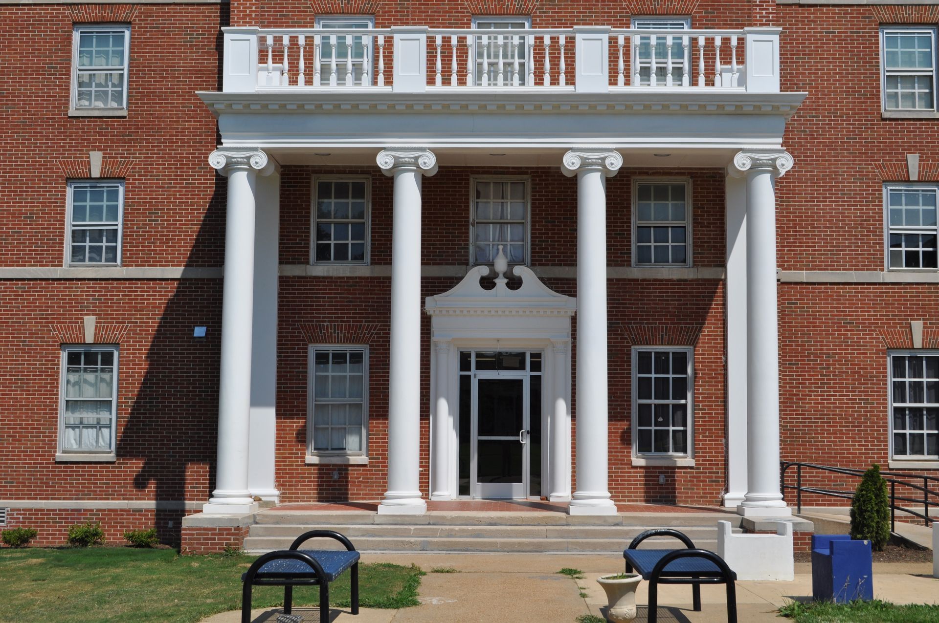 A red brick building with four white columns supporting a balcony over a central entrance door, flanked by blue benches.