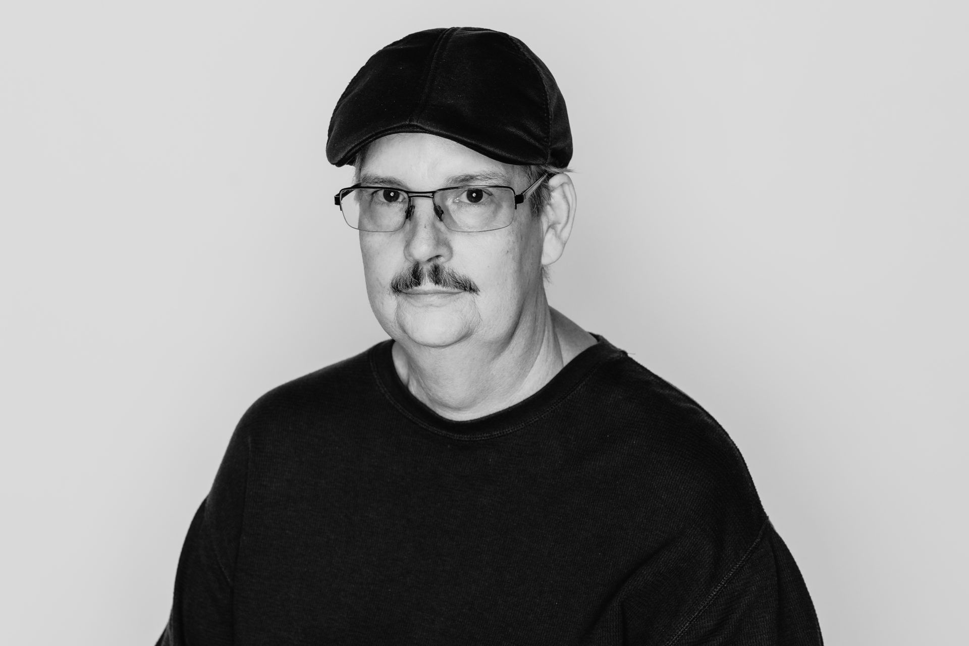 A black-and-white portrait of a person with a mustache, wearing glasses and a cap, against a light gray background.