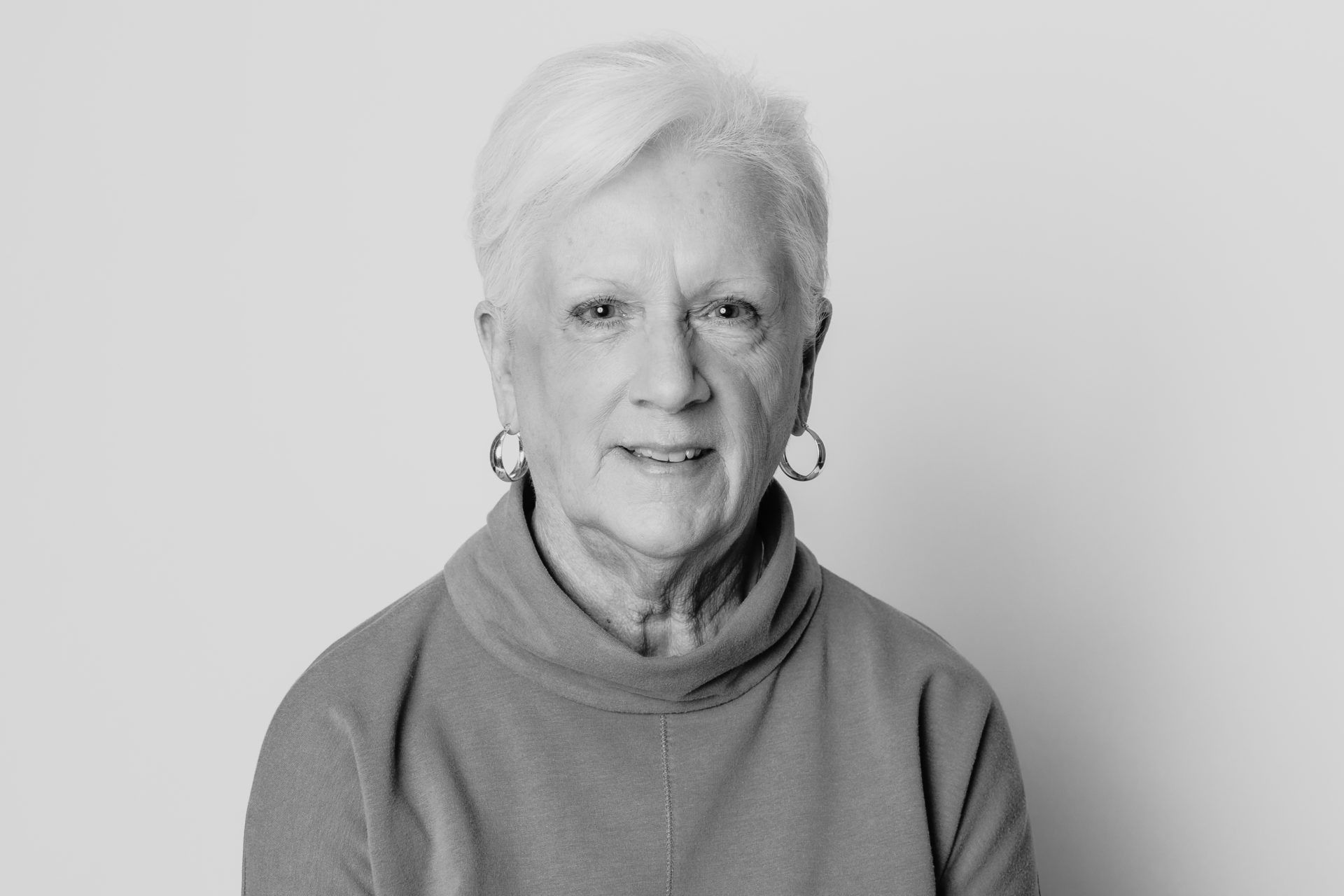 A black-and-white headshot of a person with short hair wearing hoop earrings and a high-neck top against a plain background.