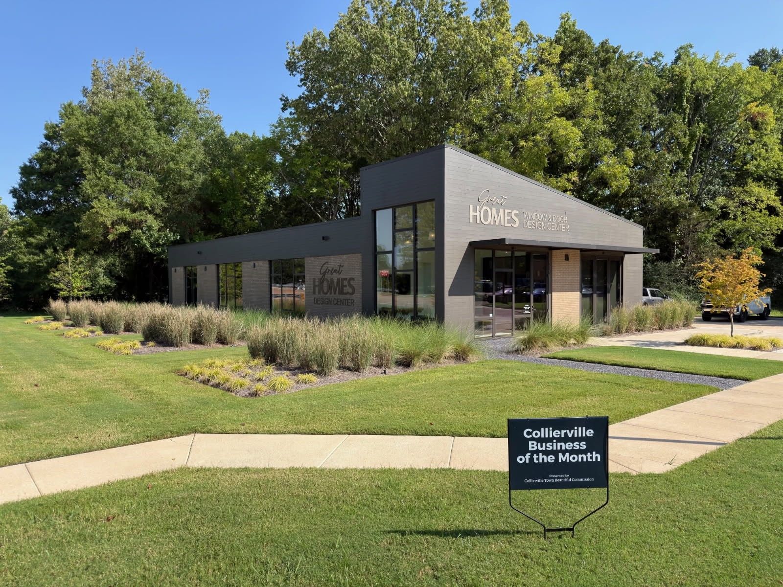 Modern gray building with a glass entrance, landscaping, and a small sign on a lawn near trees