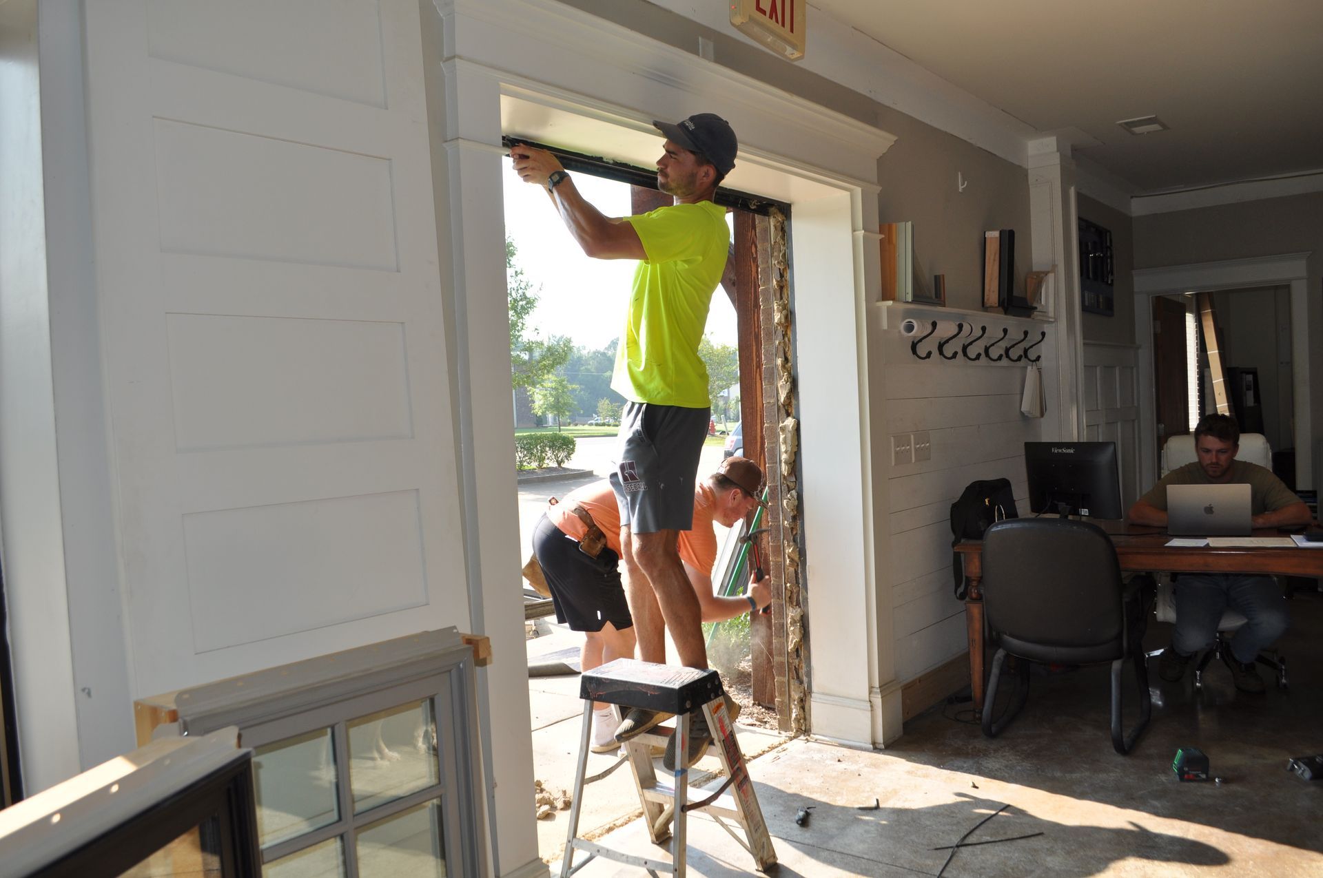 Two people work to install a door frame in a room while a third person works at a desk in the background.