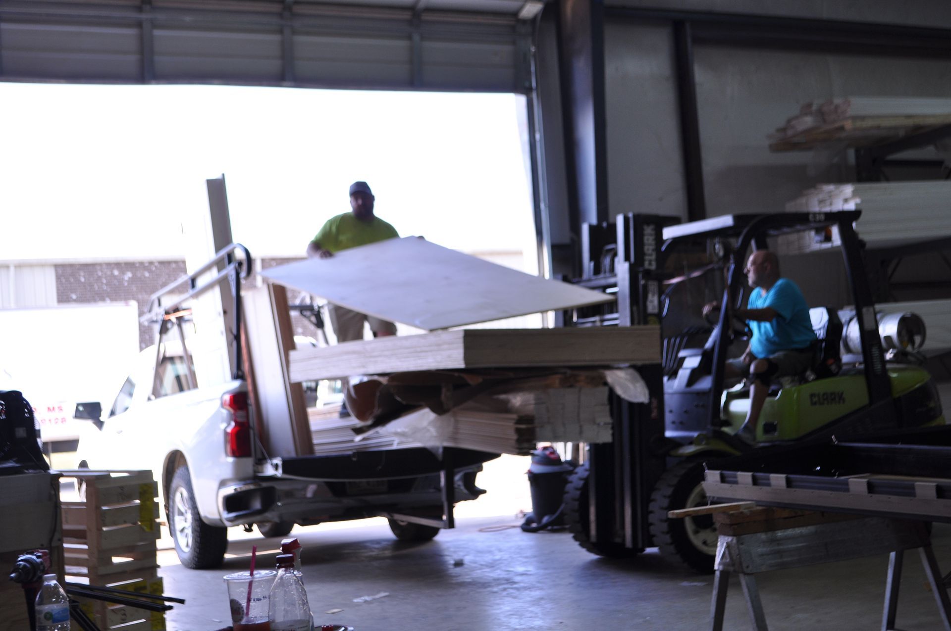 A forklift operator unloads a stack of plywood from the back of a white pickup truck inside a warehouse.