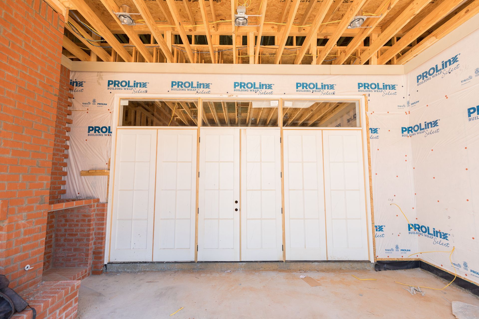A construction site interior featuring a large white door set and unfinished wood-framed ceilings and brick walls.