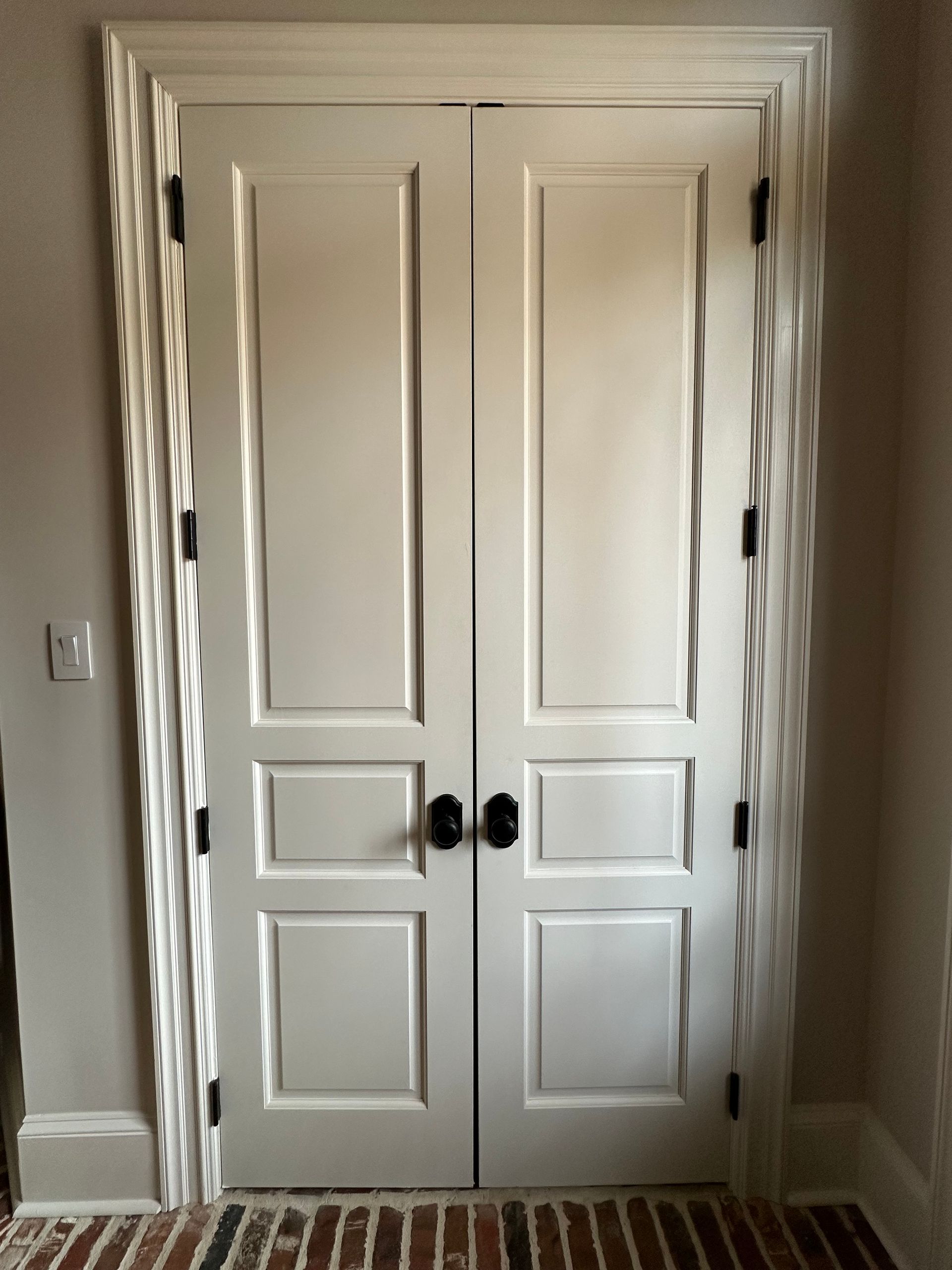 Double white paneled doors with black hardware, set in a decorative white frame against a gray wall over a brick floor.
