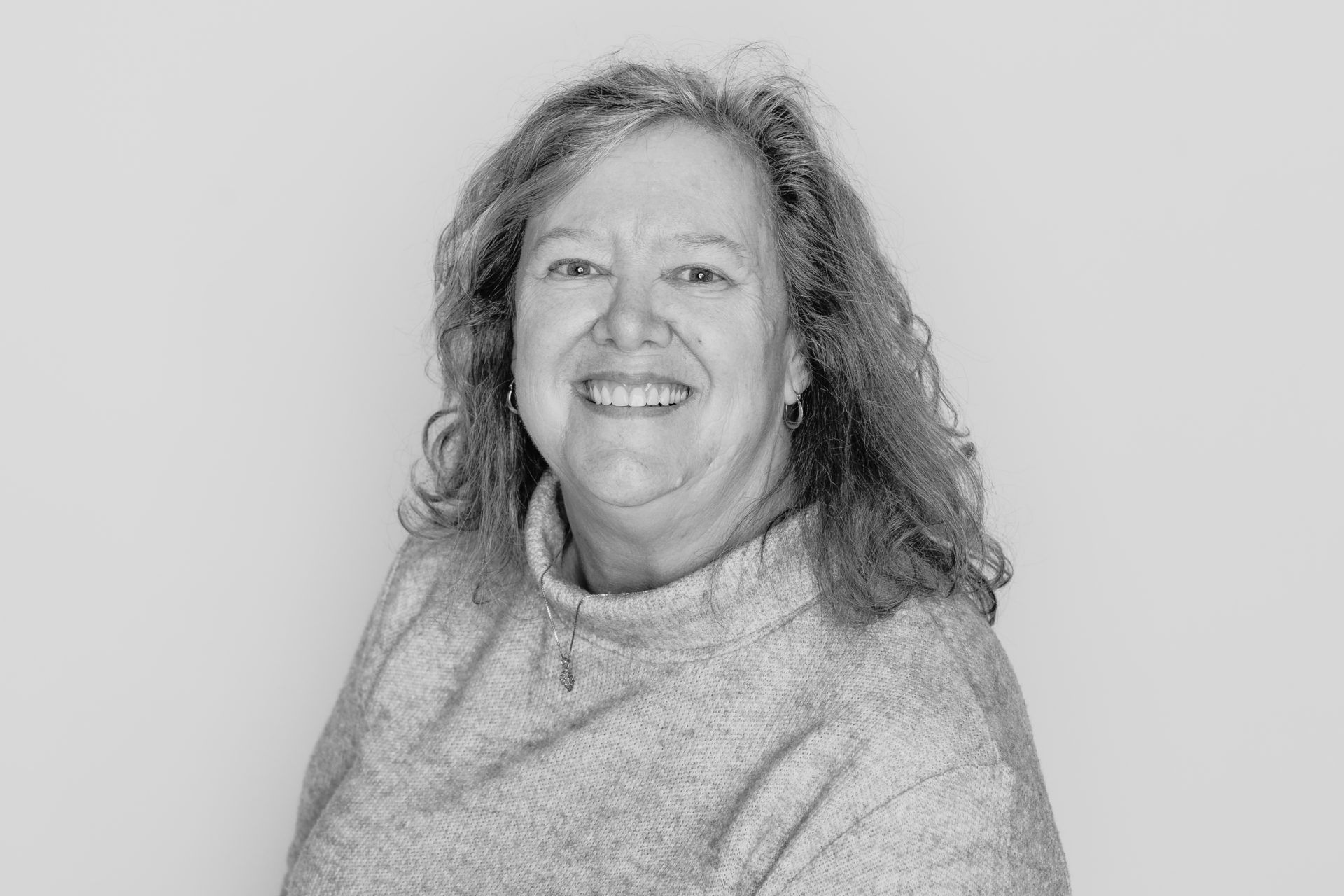 A smiling person with wavy hair, wearing a textured mock-neck sweater against a plain, light-colored background.