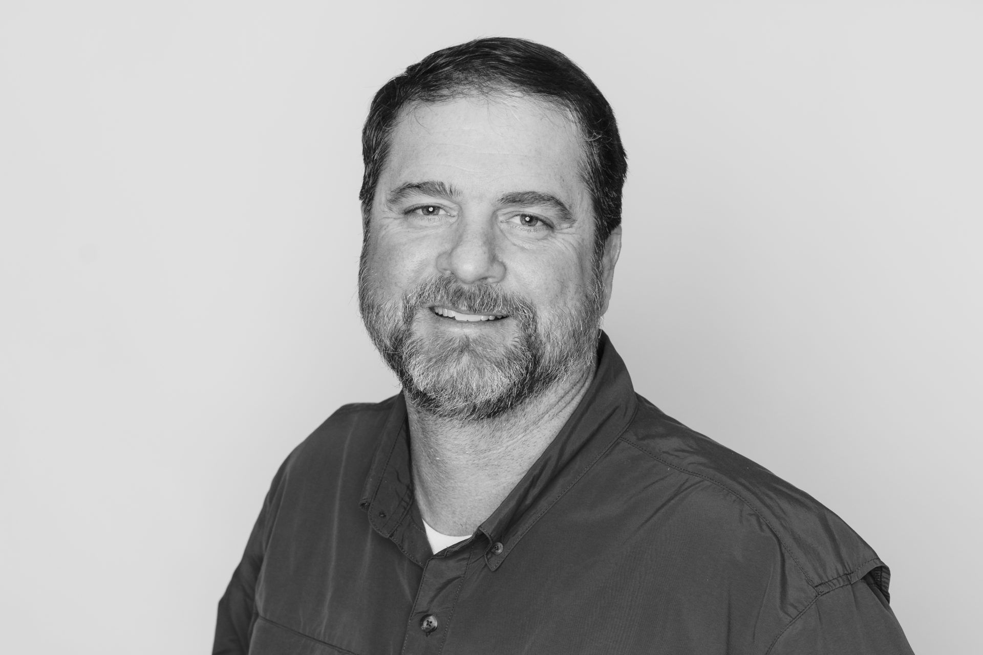 A person with a beard and short dark hair wearing a collared shirt, smiling against a plain light background.