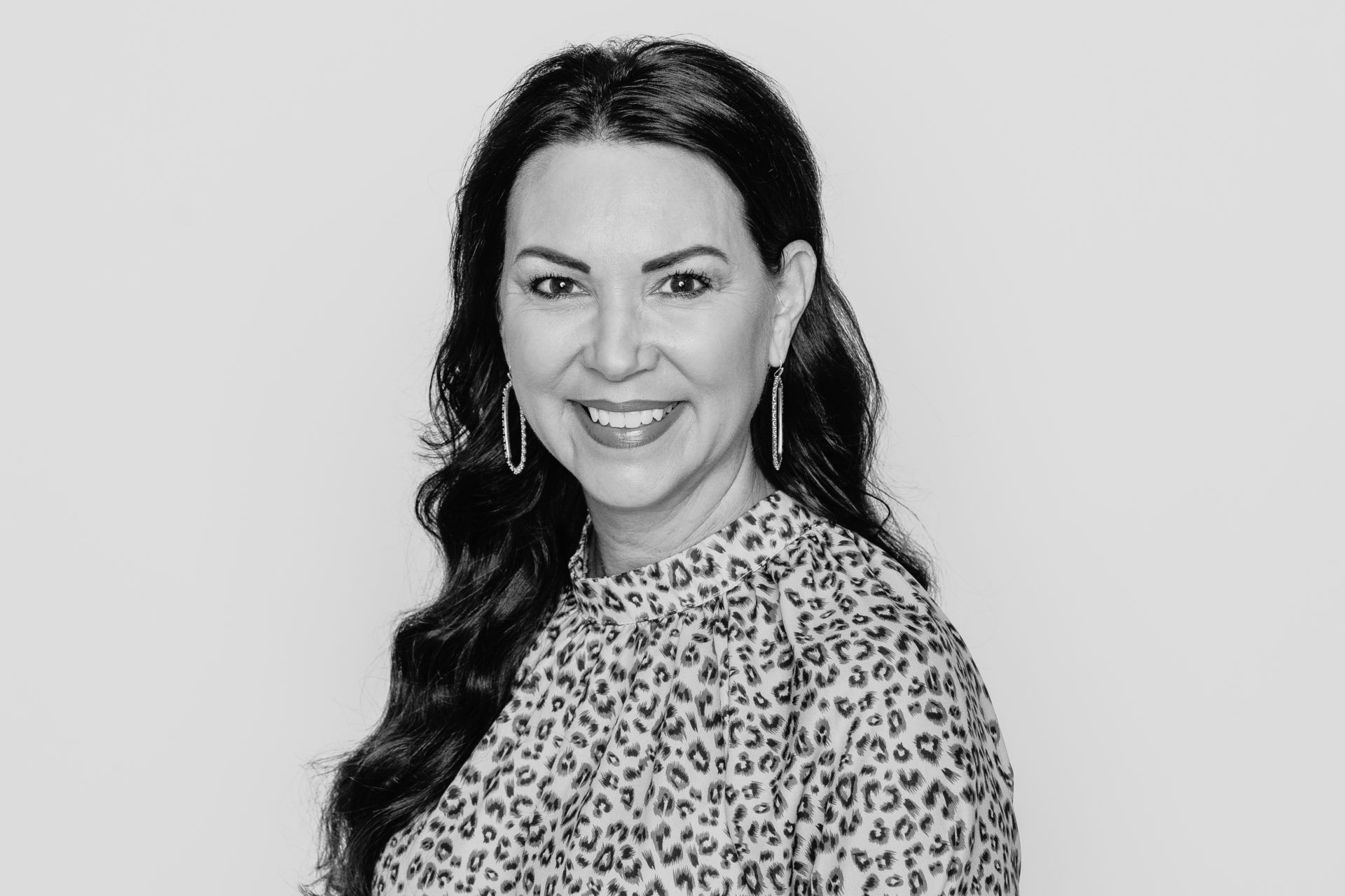 A smiling person with long, dark, wavy hair wearing a patterned top and drop earrings, captured in a black-and-white headshot.