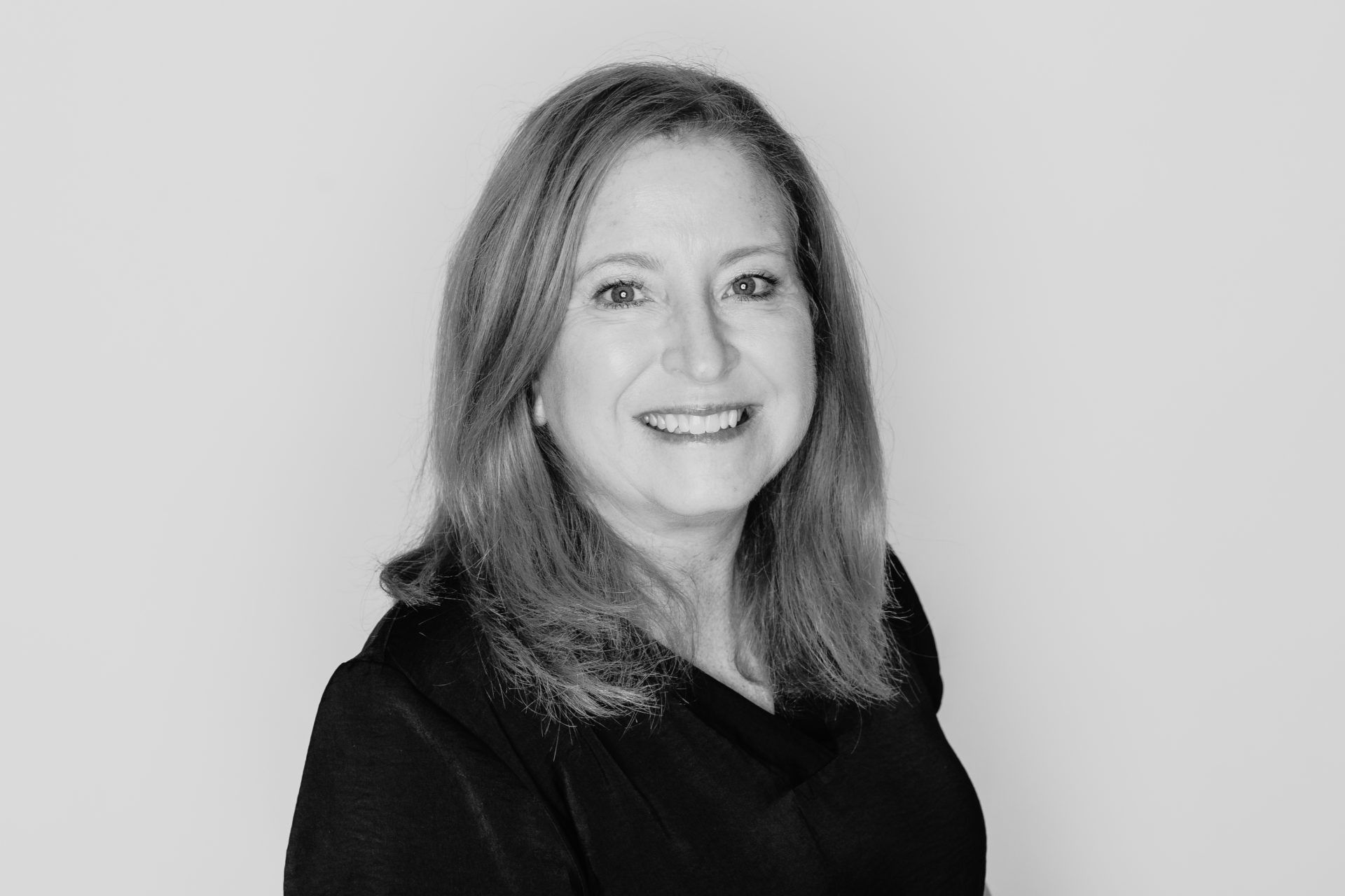 A smiling person with shoulder-length hair wearing a dark shirt against a plain, light background.
