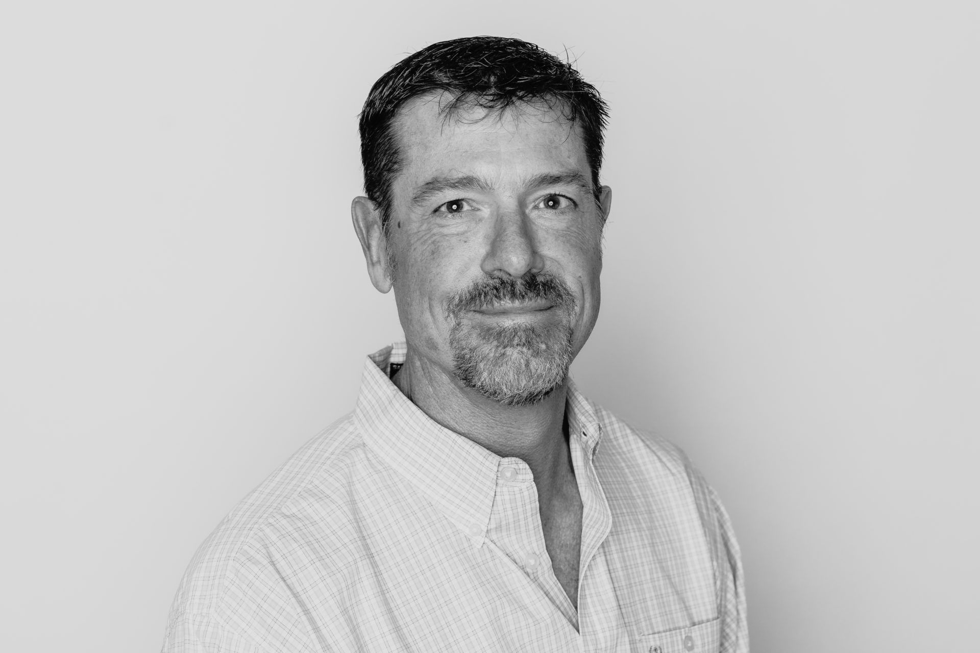 Headshot of a man with short, dark hair and facial hair, wearing a button-down shirt against a plain, light background.