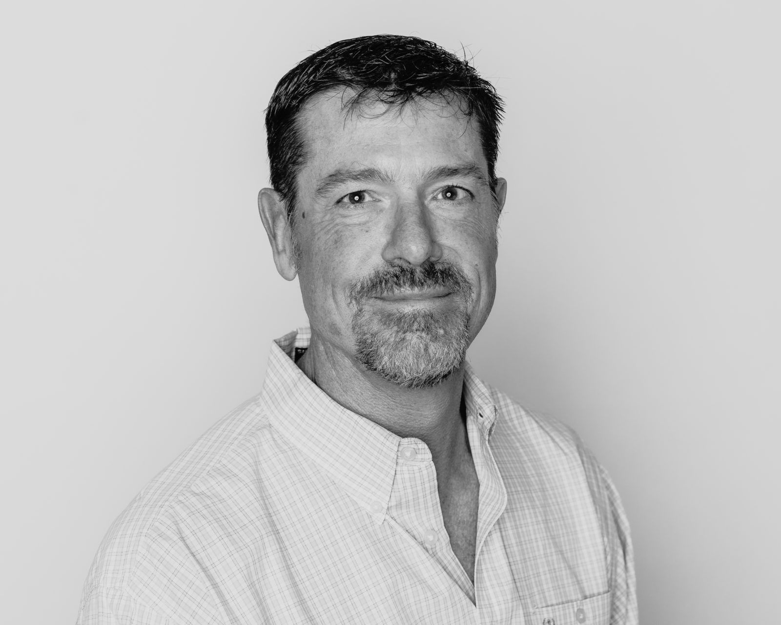Headshot of a man with short, dark hair and facial hair, wearing a button-down shirt against a plain, light background.