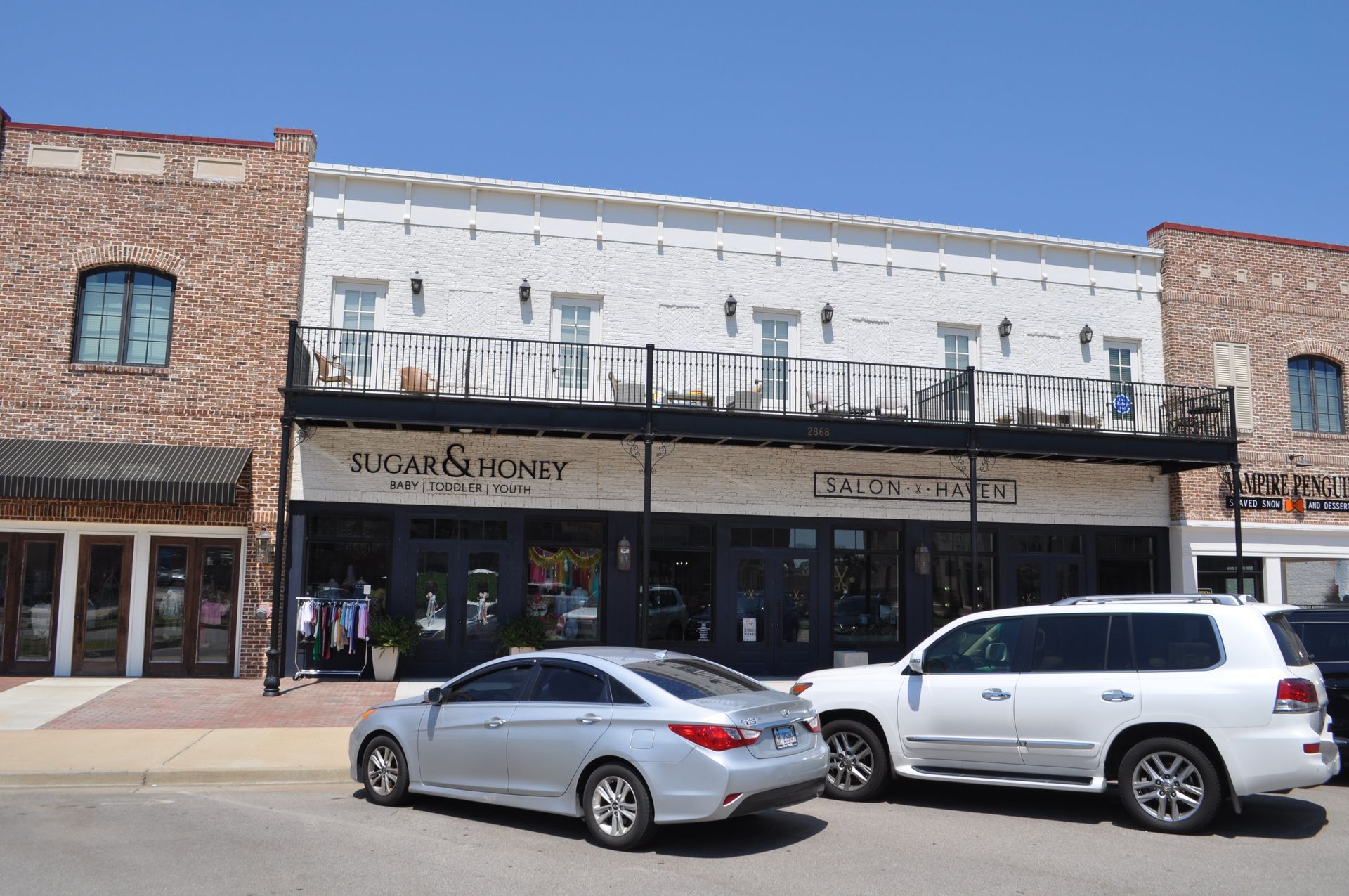 A white historic building with a balcony above storefronts, next to brick buildings, on a sunny street with two cars.