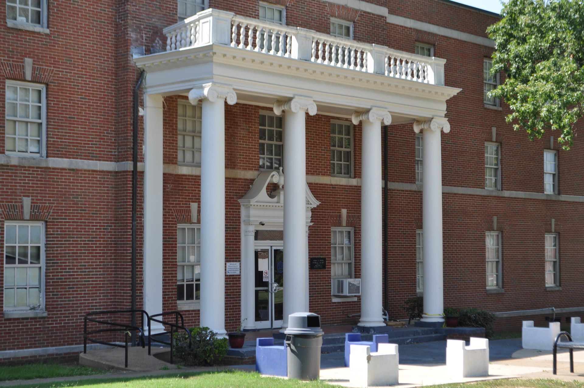 A brick building with a white columned portico entrance, a balcony railing on top, and multiple windows.
