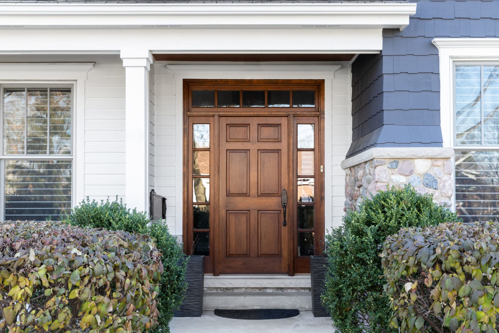 Front entrance with wooden door, white trim, stone steps, and hedges on either side.