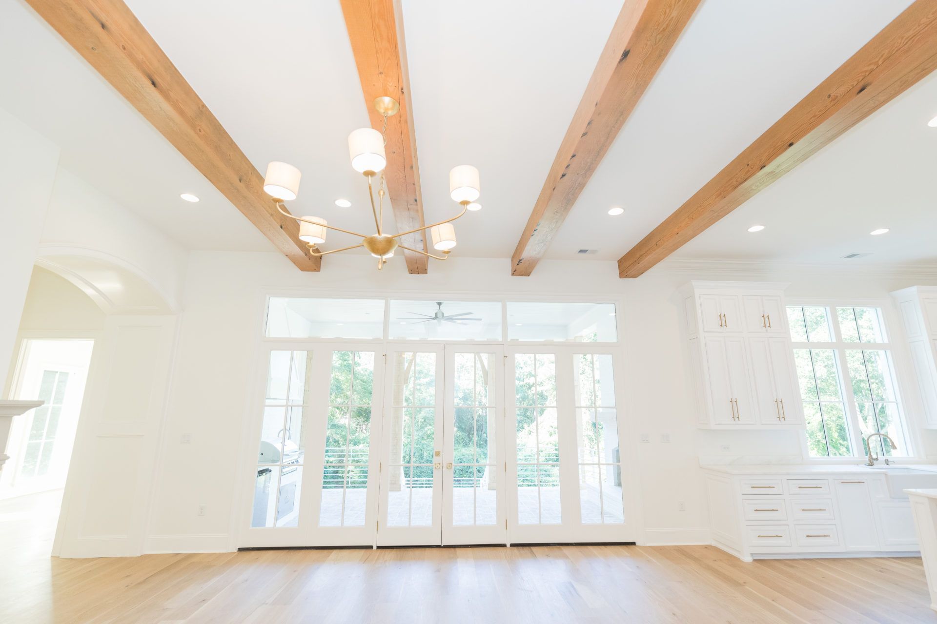 Bright empty living room with exposed wood beams, white walls, large windows, and light wood floors