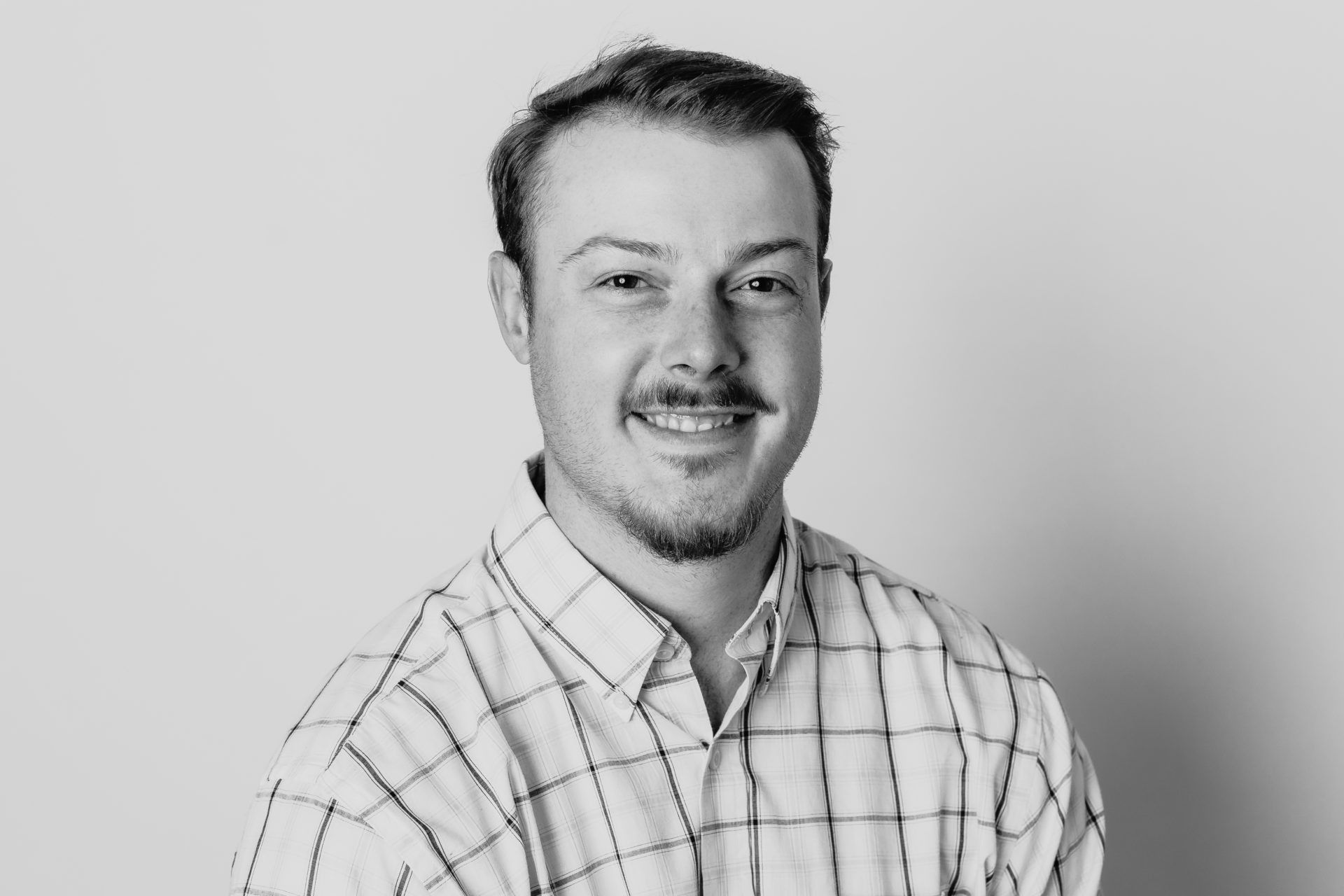A person with a beard, wearing a collared button-down shirt with a grid pattern, smiling against a light background.