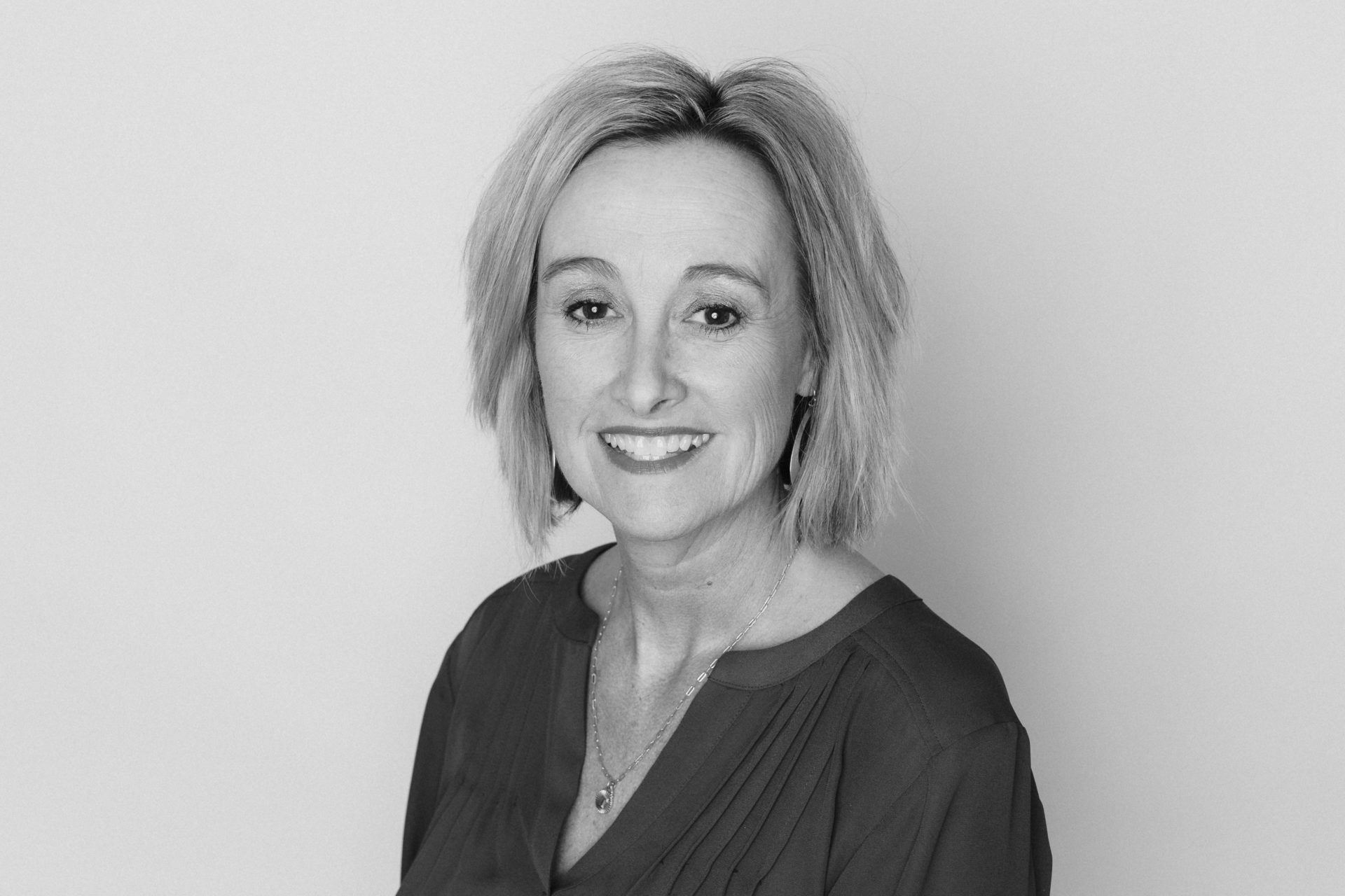 A professional headshot of a smiling woman with shoulder-length hair, wearing a dark V-neck top against a plain background.