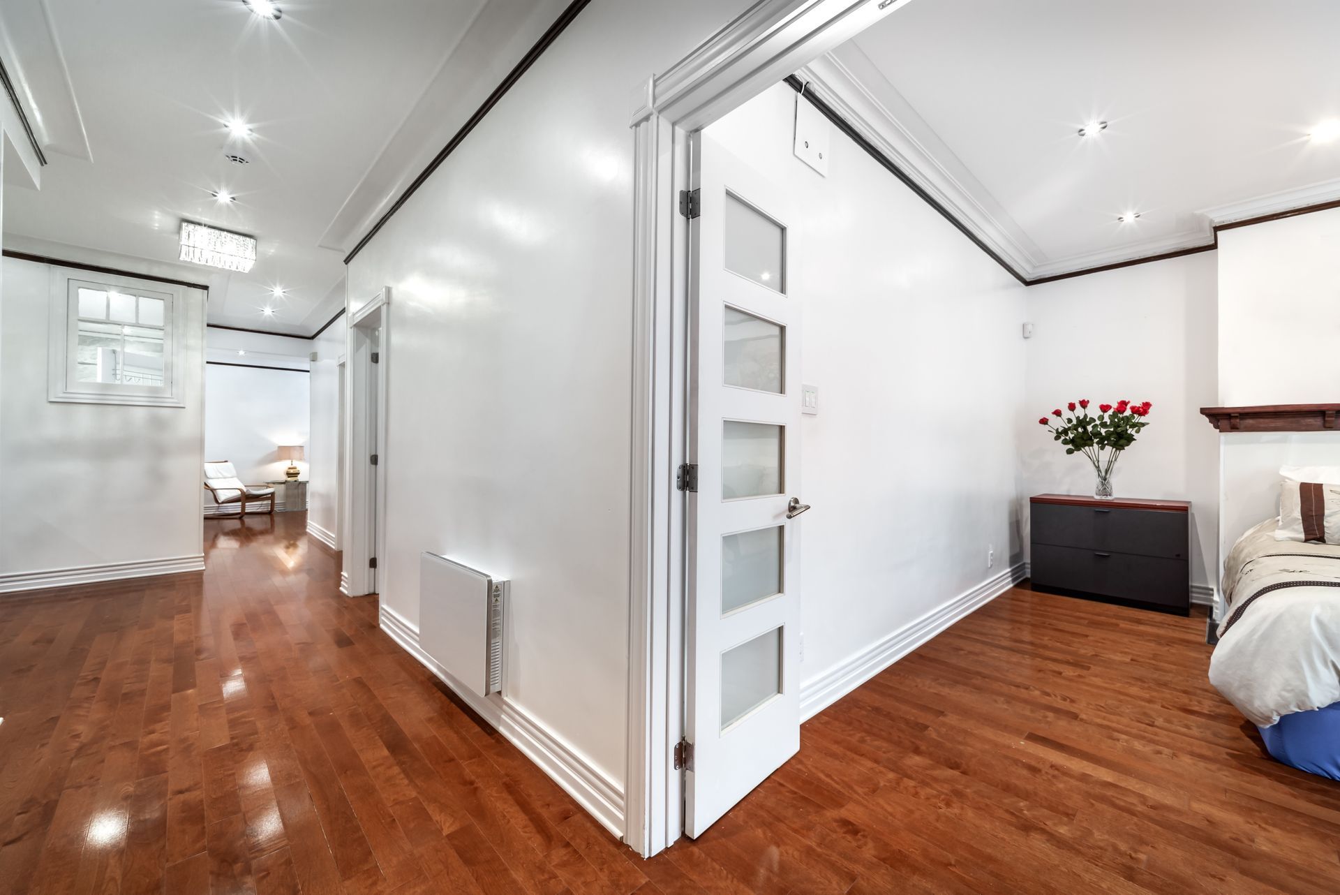 Hallway with hardwood floors, white walls, and open doors; a bed and roses are visible.