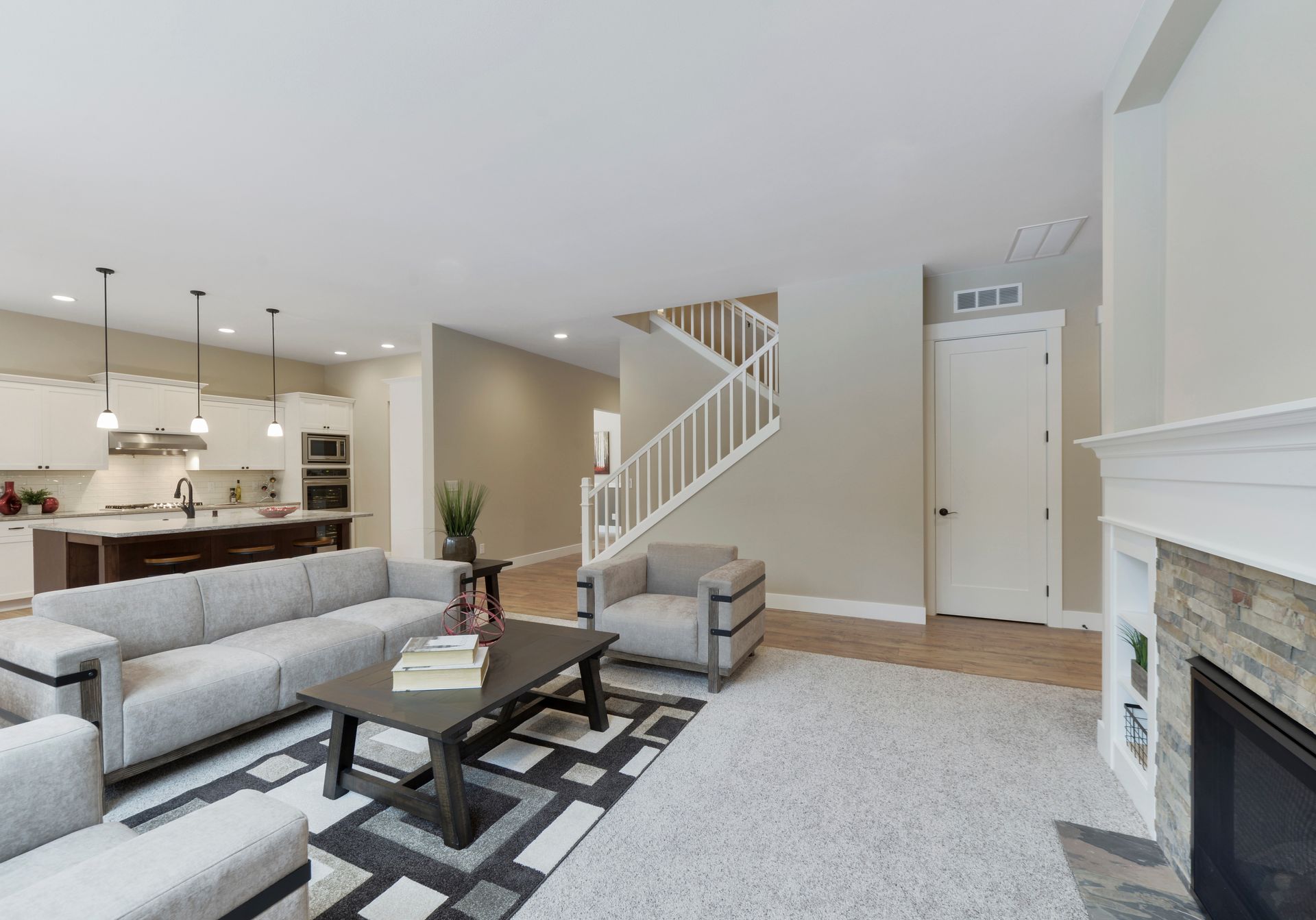 Spacious, neutral-toned living room with a kitchen, fireplace, seating area, and staircase.