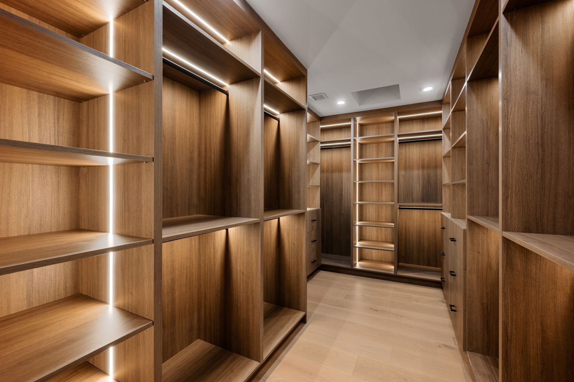 Empty walk-in closet with built-in wooden shelves and recessed lighting. Light wood flooring.