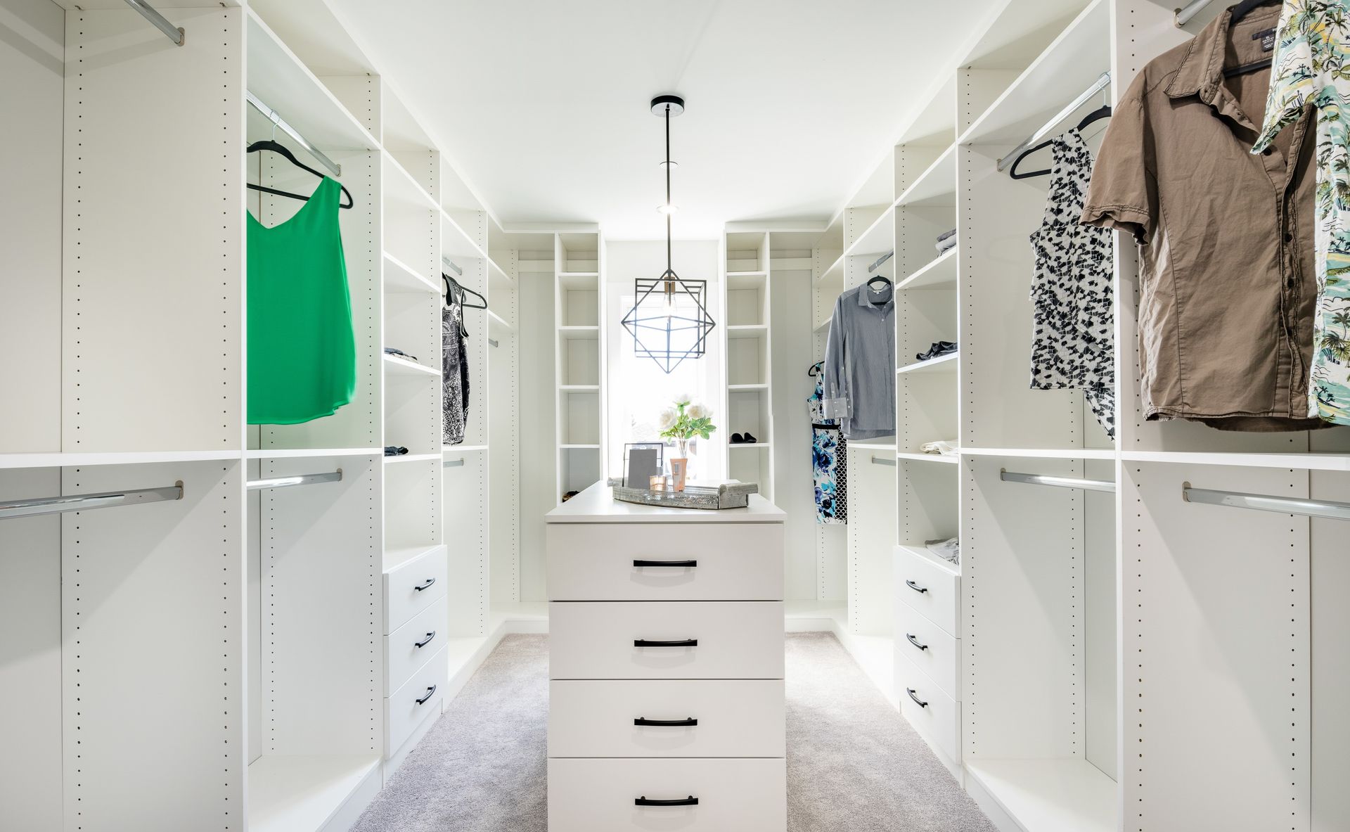 Walk-in closet with white shelves and drawers, clothes hanging. Center island with drawers, window in the back.