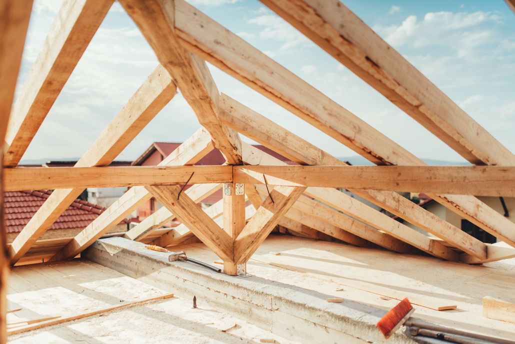 Wooden roof trusses under construction, with a partial view of the sky and surrounding houses.