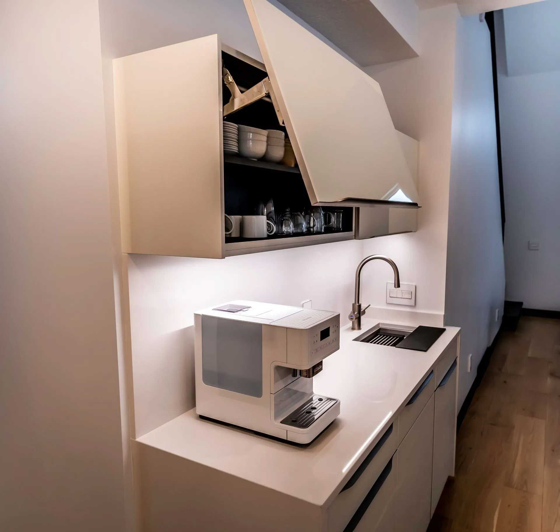 Compact, modern white kitchenette with coffee maker, sink, and overhead storage.