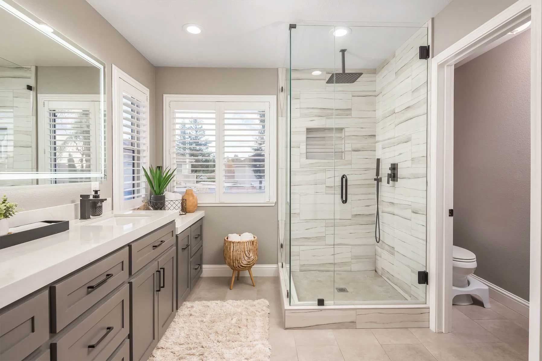 Modern bathroom with a glass shower, gray cabinets, and a window with shutters.