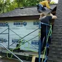 Two people on a roof working, with scaffolding holding a roll of house wrap.