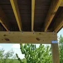 Wooden deck underside with joists, nails, and support post, with green trees in the background.