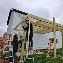 Construction workers building a wooden pergola attached to a white house, cloudy sky.