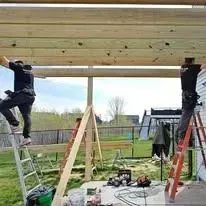 Two people building a wooden structure. One on ladder, the other on beams, sunny outdoors.