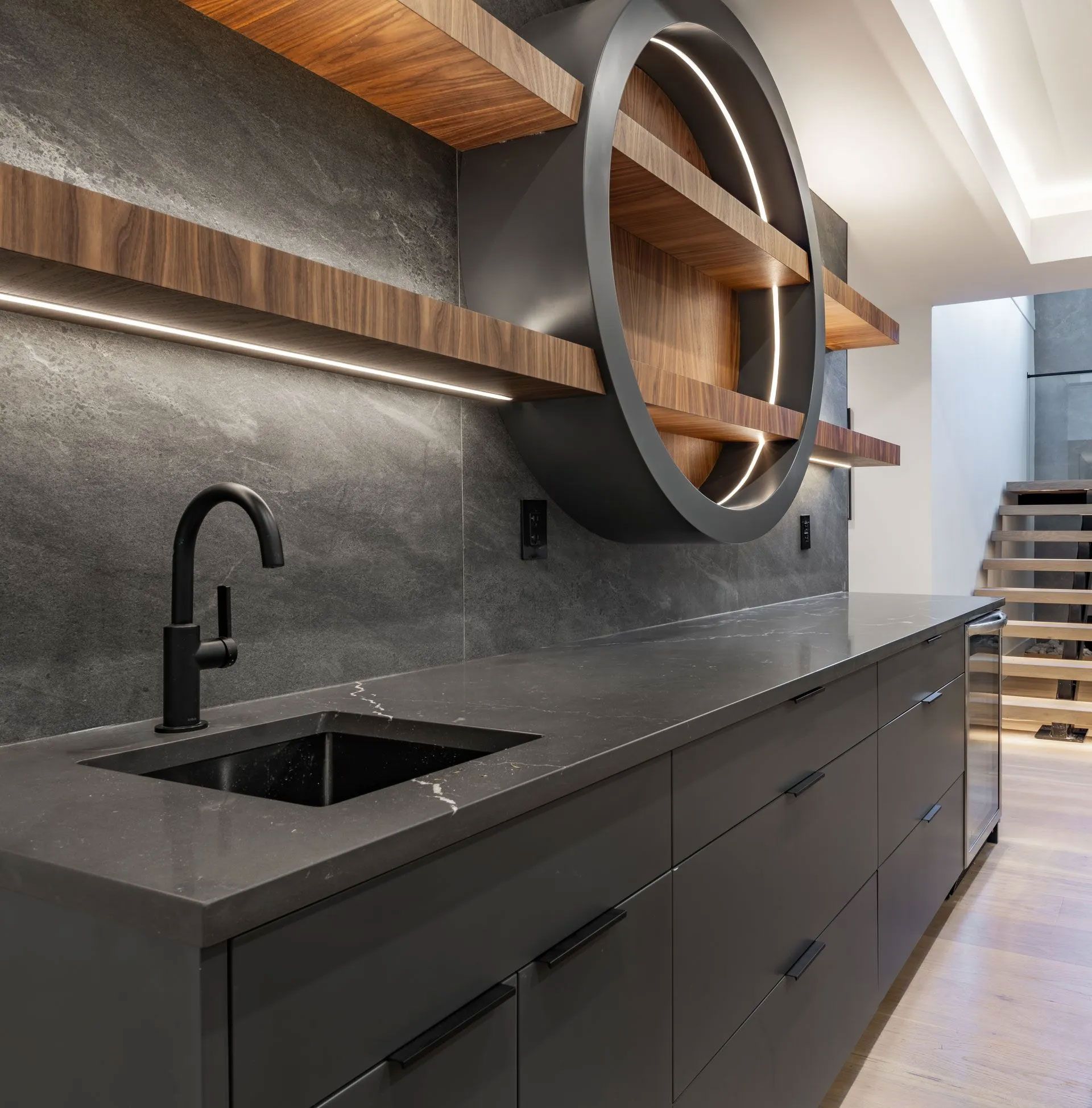Dark gray kitchen area with a black faucet, sink, countertops, and cabinets. Wooden shelves and a circular feature.