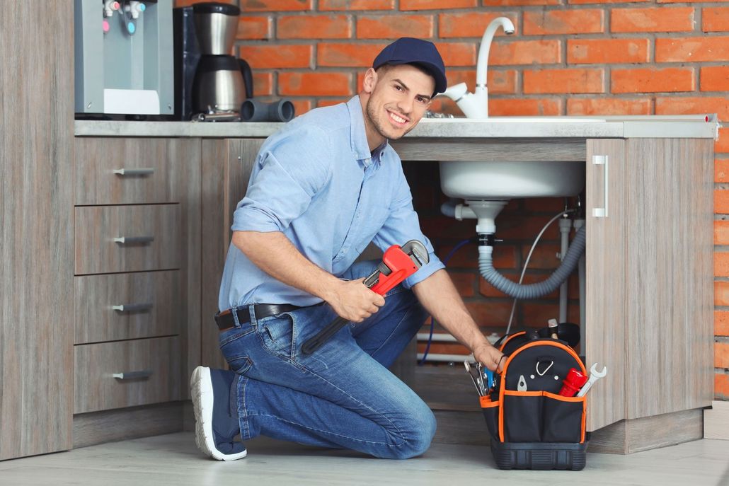 Plumber kneeling under a kitchen sink, holding a wrench, smiling. Tool bag open beside him.