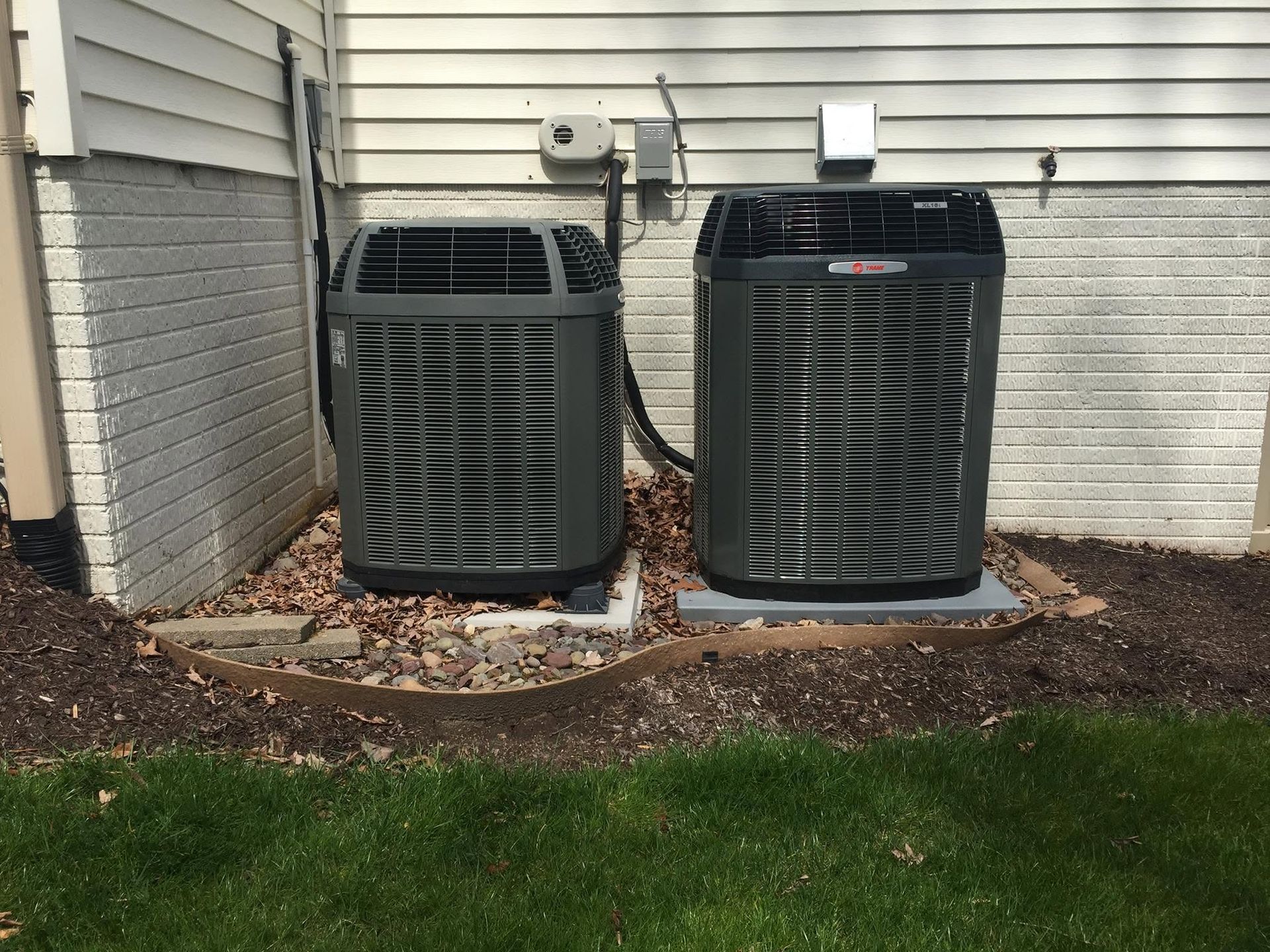 Two outdoor HVAC units beside a house, with one gray and one black, on a gravel border near grass.