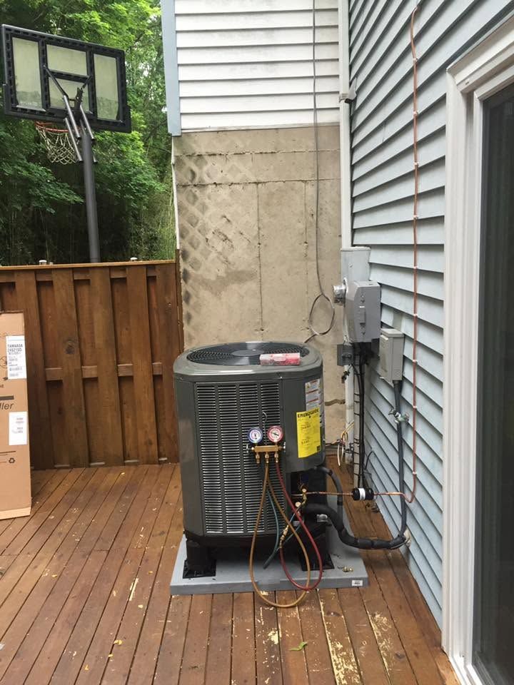 Outdoor air conditioner unit on a wooden deck beside a house, with fenced yard and basketball hoop.