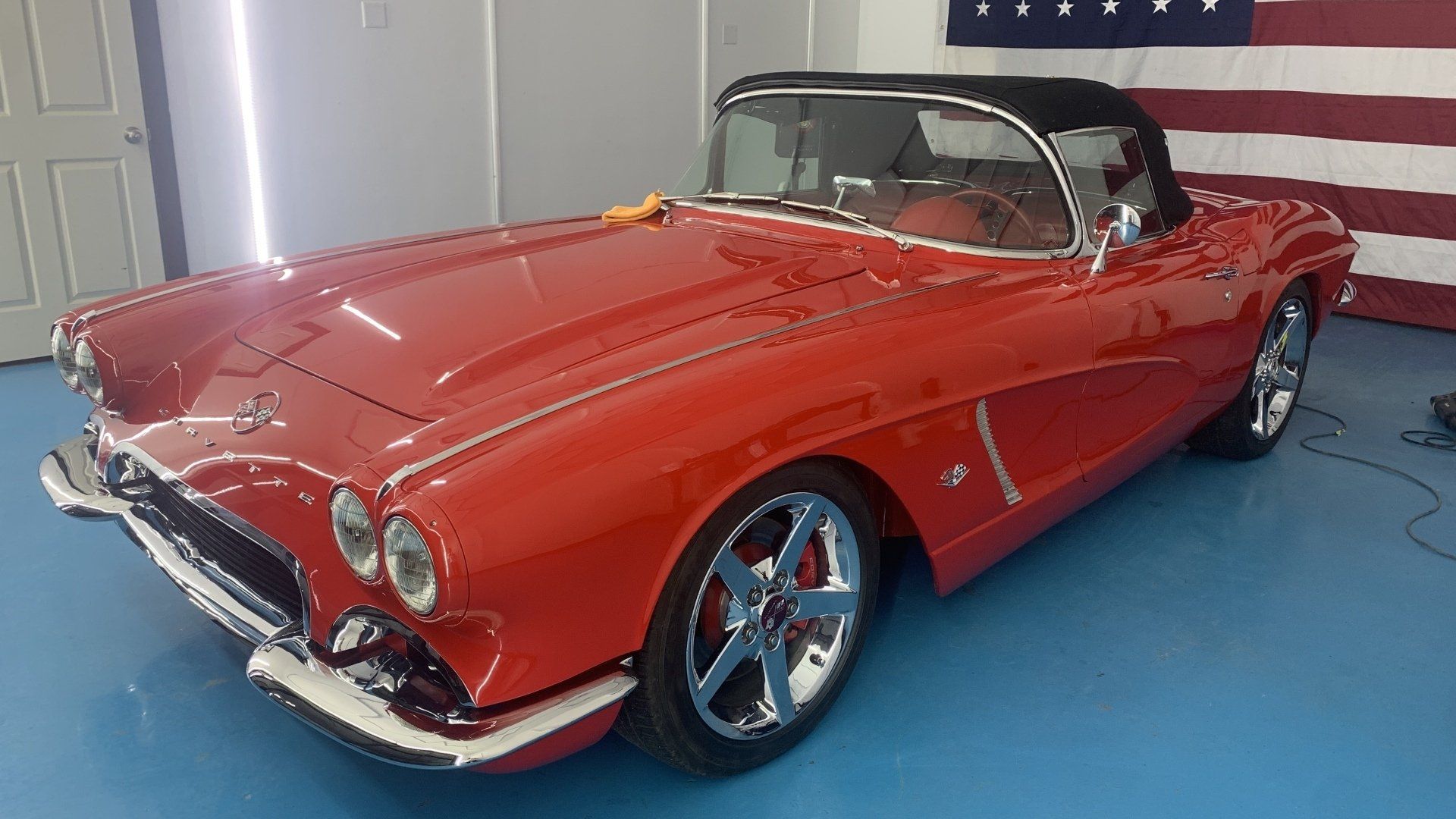 A red corvette convertible is parked in a garage next to an american flag.