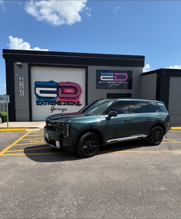 Dark green SUV parked outside a detail shop with black-and-pink signage under a blue sky