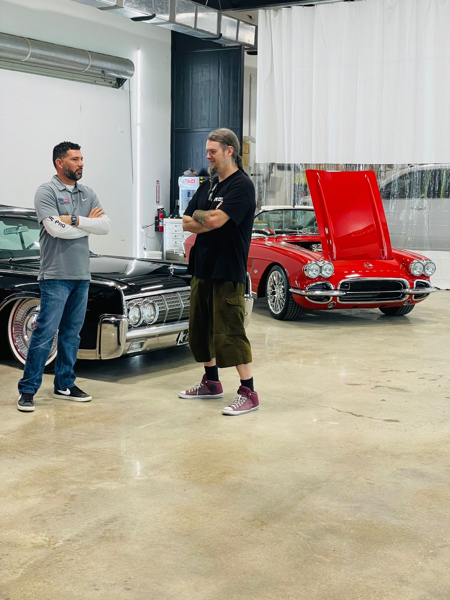 Two men are standing in front of two cars in a garage.