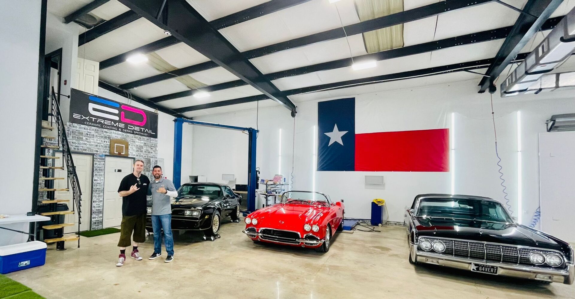 Two men are standing in a garage with cars and a texas flag.