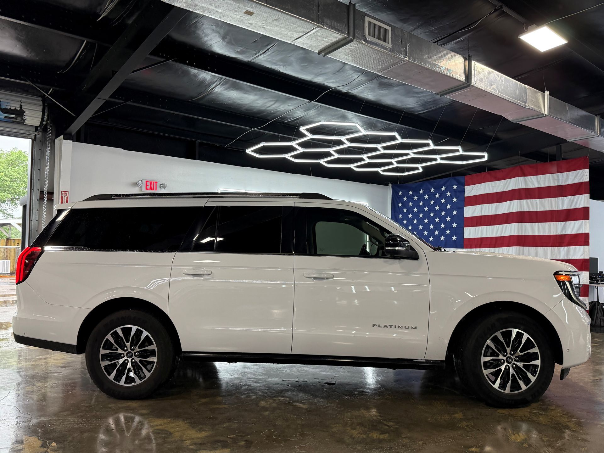 White SUV parked indoors beneath geometric lights, with a large American flag on the wall.