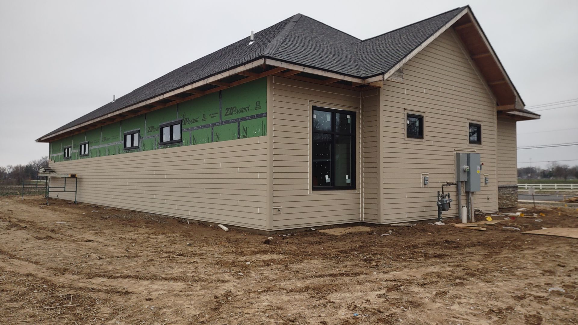 New construction building with beige siding, black windows, green sheathing, brown roof, and a dirt yard.