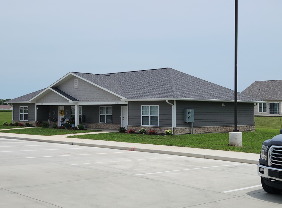 Gray and white building with a dark gray roof, surrounded by grass and a parking lot.